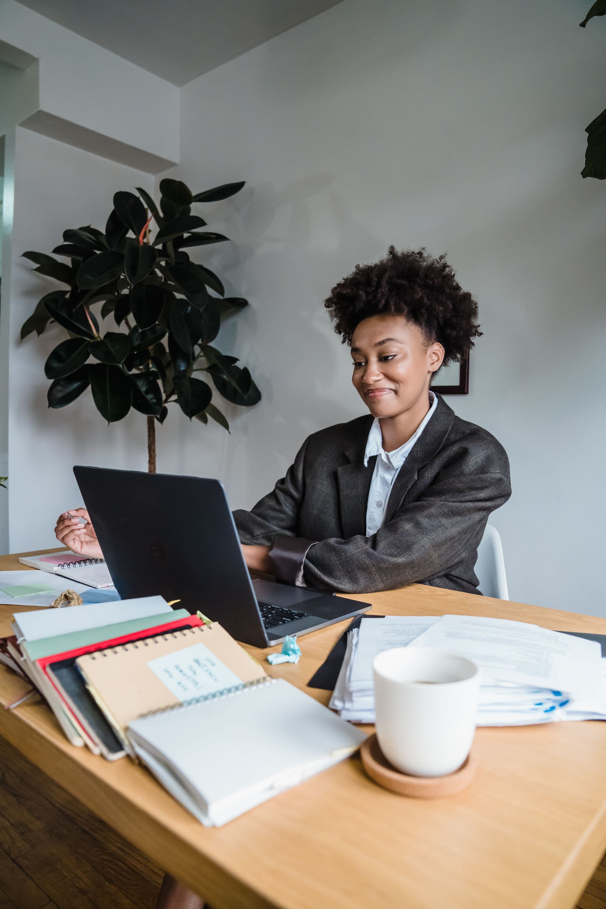 Professional in a blazer smiling while working on a laptop at a wooden desk